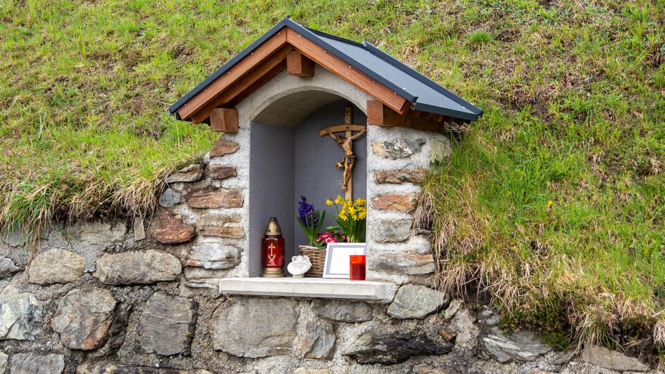 Eine kleine Nische in einer Steinmauer mit einem Holzdach. Darin befinden sich Figuren, Kerzen und Blumen zur Andacht. | © Bernhard Gruber – Photography