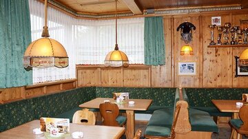 Eine gemütliche Gaststube mit holzgetäfelten Wänden und grünen Vorhängen. Die Tische sind aus Holz und mit einladenden Sitzbänken ausgestattet.
