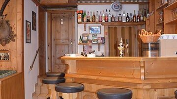 Eine gemütliche Bar mit einer Holztheke und Hockern. Im Hintergrund sind Regale mit Getränken und Gläsern zu sehen.