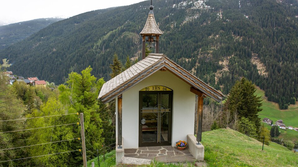 Eine kleine Kapelle steht auf einem Hügel, umgeben von Bäumen und Bergen. Im Hintergrund ist eine malerische Berglandschaft zu sehen. | © Bernhard Gruber – Photography