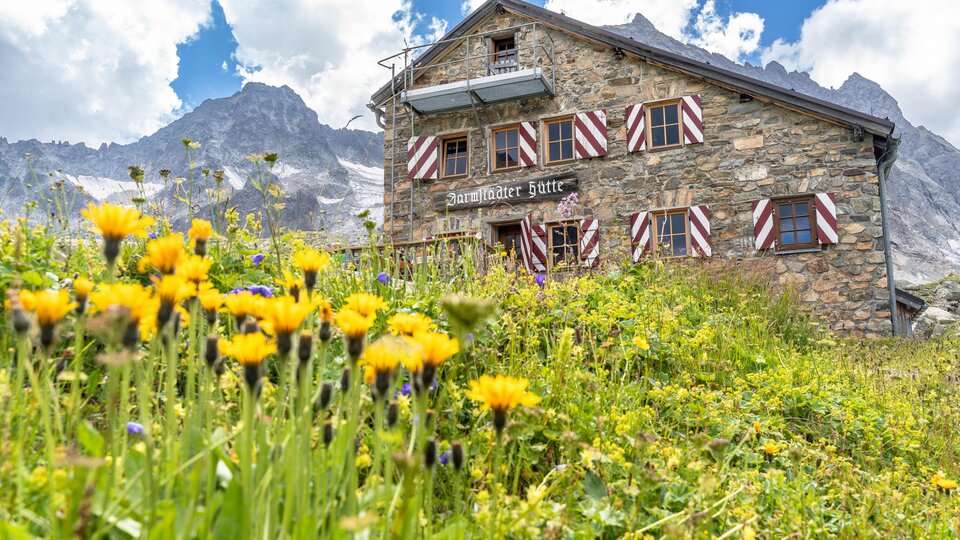 Eine malerische Berghütte aus Stein mit bunten Fensterläden. Der Garten ist mit gelben Blumen und einer beeindruckenden Bergkulisse umgeben.