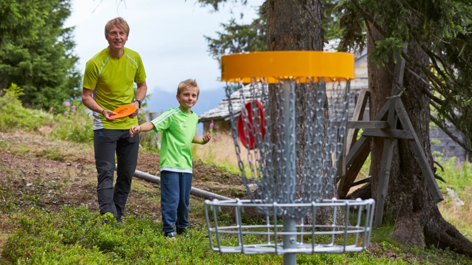 Ein Mann und ein Junge stehen in einem Wald und spielen Disc Golf. Im Hintergrund ist der Korb für das Spiel zu sehen.
