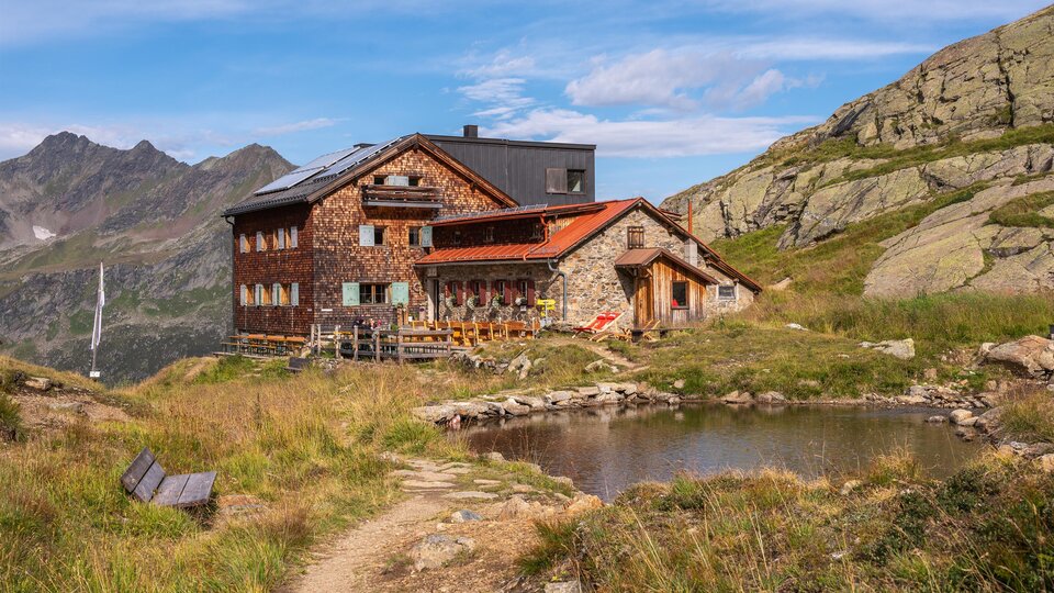 Eine malerische Berghütte inmitten einer grünen Landschaft. Im Vordergrund befindet sich ein kleiner Teich und im Hintergrund sind beeindruckende Berge zu sehen.