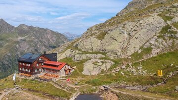 Eine Berghütte liegt malerisch in einer gebirgigen Landschaft. Im Hintergrund sind hohe Berge und ein klarer Himmel zu sehen.