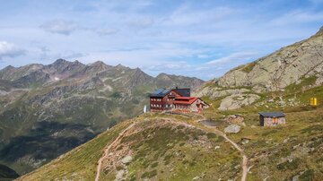 Eine beeindruckende Berglandschaft mit einer Berghütte auf einem Gipfel. Der Himmel ist teilweise bewölkt und die umliegenden Berge sind gut sichtbar.