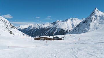 Eine malerische Winterlandschaft mit schneebedeckten Bergen. Im Vordergrund sind Skipisten und eine gemütliche Berghütte zu sehen.