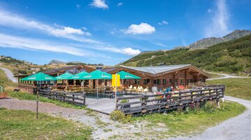 Eine gemütliche Berghütte mit Terrasse und Sonnenschirmen. Umgeben von grünen Wiesen und Bergen unter einem blauen Himmel.
