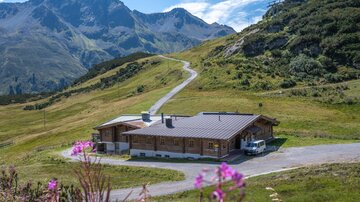 Ein modernes Holzhaus in einer malerischen Berglandschaft. Umgeben von grünen Wiesen und hohen Bergen, bietet die Umgebung eine ruhige und entspannende Atmosphäre.