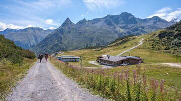 Ein malerischer Weg führt durch eine grüne Landschaft mit Bergen im Hintergrund. Wanderer gehen auf den Pfad zur nächsten Hütte.