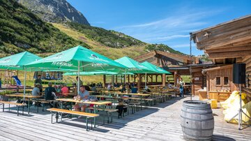 Eine schöne Außenterrasse mit Tischen und grünen Sonnenschirmen in den Bergen. Die Umgebung ist grün und malerisch, ideal für ein entspanntes Essen im Freien.