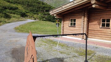 Ein rustikales Holzhaus in einer bergigen Landschaft. Im Vordergrund steht ein Metallschild, das den Eingang markiert.