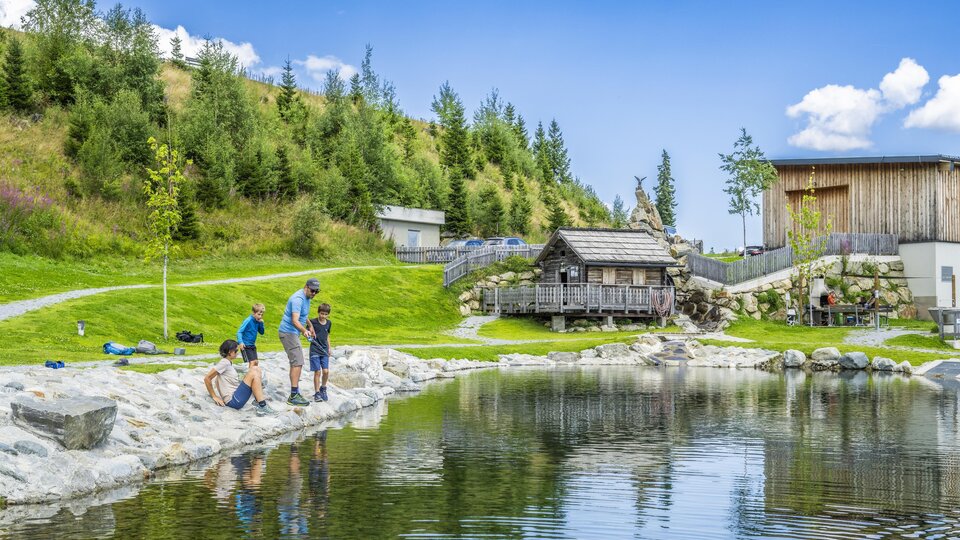 Ein ruhiger See umgeben von grünen Wiesen und Bäumen. Kinder und Erwachsene genießen die Natur und verbringen Zeit am Ufer.