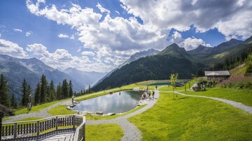 Eine malerische Landschaft mit einem klaren Teich und grünen Wiesen. Im Hintergrund sind majestätische Berge und ein blauer Himmel mit weißen Wolken zu sehen.