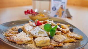 Ein Teller mit süßen Pfannkuchen, bestreut mit Puderzucker und verziert mit Beeren und Minze. Dazu gibt es eine Schale mit Fruchtsoße.