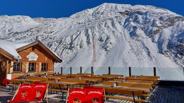 Eine Berghütte mit Holzterrasse und mehreren Tischen und Stühlen steht vor einer beeindruckenden Bergkulisse. Der Schnee bedeckt die Landschaft unter einem klaren blauen Himmel.