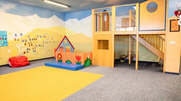 Ein helles und freundliches Spielzimmer mit bunten Wänden. Es gibt einen Spielbereich mit einer kleinen Rutsche und einen gemütlichen Sitzbereich.