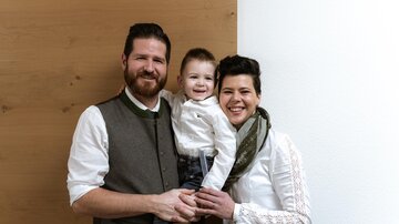 Eine Familie mit einem kleinen Kind steht zusammen vor einer hellen Wand. Alle tragen elegante, lässige Kleidung und lächeln freundlich.
