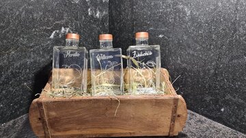 Drei Glasflaschen in einem Holzbehälter, umgeben von Heu. Die Flaschen sind mit Korken verschlossen und beschriftet.