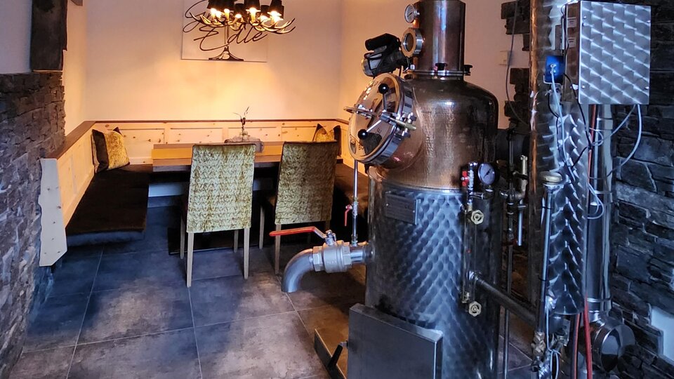 Ein gemütlicher Raum mit einem großen Destilliergerät. Im Hintergrund stehen mehrere Stühle an einem Tisch.