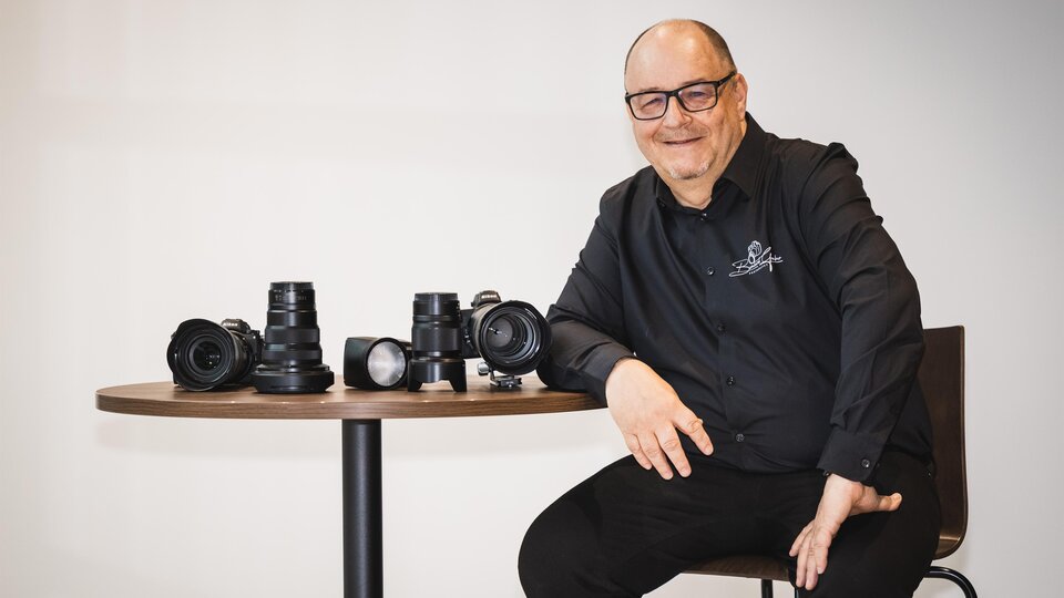 Ein lächelnder Mann sitzt an einem Tisch und präsentiert mehrere Kameras und Objektive. Er trägt ein schwarzes Hemd und wirkt freundlich und kompetent. | © Bernhard Gruber – Photography