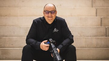 Ein Fotograf sitzt auf einer Treppe und hält einen Kamerabody in der Hand. Er trägt ein schwarzes Hemd und lächelt in die Kamera. | © Bernhard Gruber – Photography