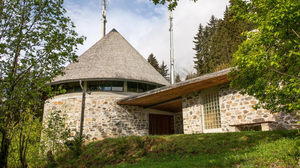 Ein modernes Gebäude in runder Form mit einem spitzen Dach und Fensterfronten. Umgeben von Bäumen und Grünflächen in einer ländlichen Umgebung. | © Bernhard Gruber – Photography