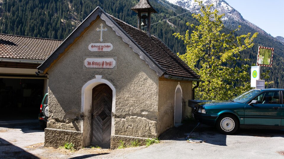 Eine kleine Kapelle mit einer eleganten Dachform und einem Kreuz. Im Hintergrund sind Berge und Bäume zu sehen. | © Bernhard Gruber – Photography