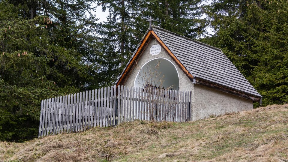 Eine kleine Hütte mit einem Holzzaun in einem Waldgebiet. Der Himmel ist bewölkt, und der Boden ist mit Gras und Laub bedeckt. | © Bernhard Gruber – Photography