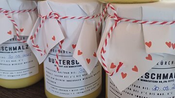 Drei Gläser mit Butterschmalz, dekoriert mit Papier und einem roten Band. Die Etiketten enthalten Informationen über das Produkt und das Haltbarkeitsdatum.