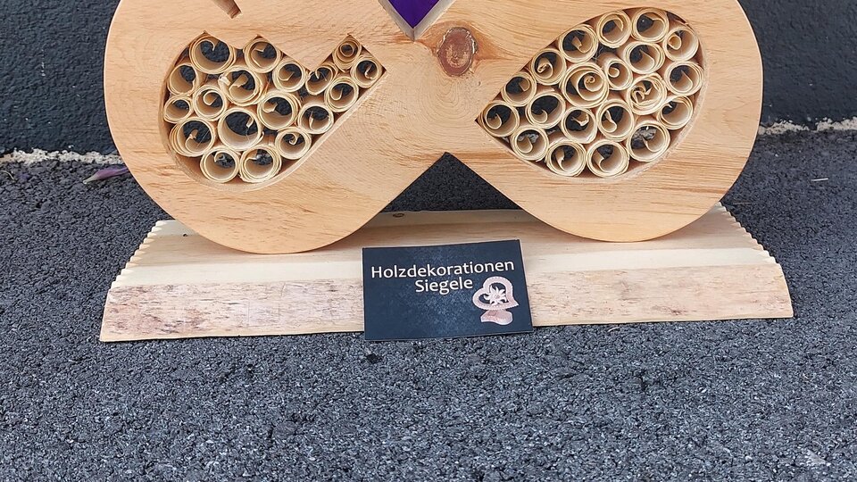 Eine dekorative Holzskulptur in Form eines Unendlichkeitssymbols mit Herzmotiven und einem spiralartigen Muster. Im Hintergrund sind lila Blumen zu sehen.
