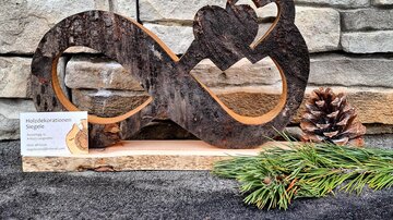 Eine Holzskulptur in Form einer Unendlichkeitsschleife mit einem Herz. Neben der Skulptur liegen ein Tannenzapfen und einige Tannennadeln.