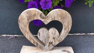 Eine Holzskulptur in Herzform mit einem kleineren Herz in der Mitte. Im Hintergrund blühen lila Petunien.