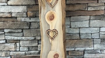 Ein dekorativer Holzblock mit zwei herzförmigen Ausschnitten. Im Hintergrund sind bunte Blumen und eine Steinmauer sichtbar.