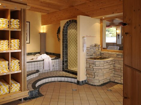 Ein entspannender Wellnessbereich mit ordentlich gestapelten Handtüchern in Regalen. Im Hintergrund sind ein Whirlpool und eine einladende Sauna zu sehen.
