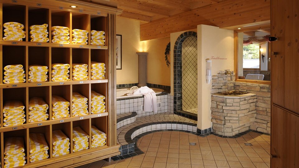 Ein entspannender Wellnessbereich mit ordentlich gestapelten Handtüchern in Regalen. Im Hintergrund sind ein Whirlpool und eine einladende Sauna zu sehen.