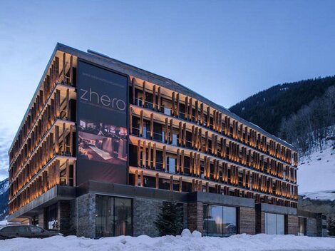 Ein modernes Hotelgebäude mit Holzverkleidung und großen Fenstern. Umgeben von schneebedeckten Bergen und einem klaren Himmel.