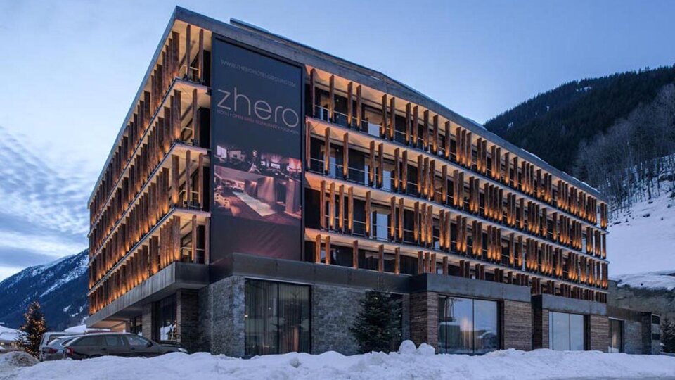 Ein modernes Hotelgebäude mit Holzverkleidung und großen Fenstern. Umgeben von schneebedeckten Bergen und einem klaren Himmel.