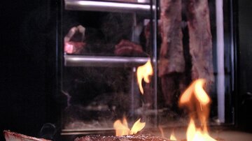 Ein saftiges Steak brutzelt auf einem Grill, während Flammen umherzuckt. Im Hintergrund sind große Fleischstücke sichtbar.