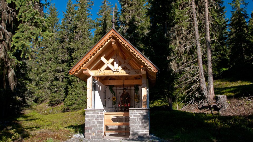 Eine kleine Holzkapelle umgeben von hohen Bäumen. Der Himmel ist blau und die Umgebung ist grün und friedlich. | © Bernhard Gruber – Photography