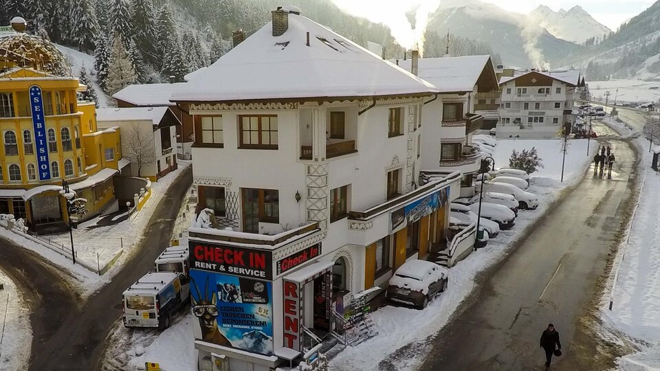 Ein charmantes Gebäude im Schnee mit einer "CHECK IN"-Anzeige. Im Hintergrund sind verschneite Berge und eine ruhige Straße zu sehen.