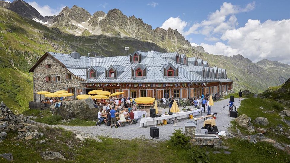 Eine gemütliche Berghütte mit Sitzgelegenheiten im Freien und vielen Gästen. Die Umgebung ist von beeindruckenden Bergen und einer klaren Bl sky umgeben.
