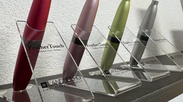 Vier verschiedene kosmetische Geräte in unterschiedlichen Farben stehen auf einem Regal. Sie sind in transparenten Halterungen platziert und präsentieren die Marke "FeatherTouch".