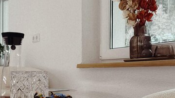 Ein modernes Wohnzimmer mit einem runden Tisch und einer dekorativen Glaskaraffe. Auf der Fensterbank stehen farbige Trockenblumen in einer Vase.