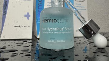Ein flüssiges Hautserum in einer Glasflasche steht im Vordergrund. Im Hintergrund sind weitere Hautpflegeprodukte in eleganter Verpackung zu sehen.