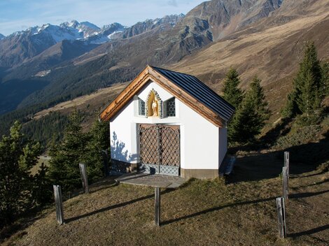 A small chapel on a hill, surrounded by mountains and forests. The landscape features clear skies and mountain peaks in the background. | © Bernhard Gruber – Photography