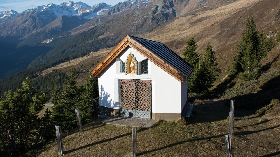 A small chapel on a hill, surrounded by mountains and forests. The landscape features clear skies and mountain peaks in the background. | © Bernhard Gruber – Photography