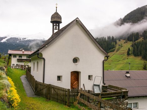 A small, white church with a bell tower stands in a picturesque landscape. In the background, green hills and cloudy sky are visible. | © Bernhard Gruber – Photography