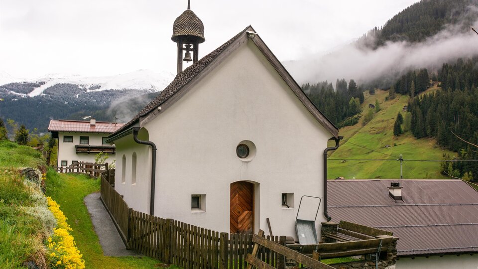 A small, white church with a bell tower stands in a picturesque landscape. In the background, green hills and cloudy sky are visible. | © Bernhard Gruber – Photography