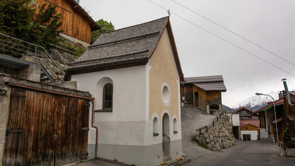A small, white chapel stands on a narrow street, surrounded by traditional wooden houses. The sky is overcast and it seems to be a quiet, rural setting. | © Bernhard Gruber – Photography