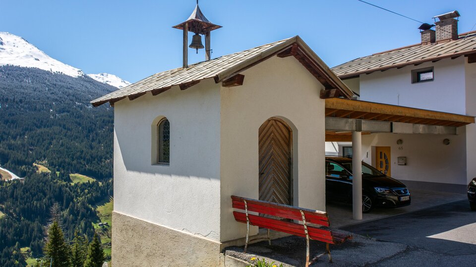 A traditional house with a small bell on the roof, surrounded by mountains. In the foreground, there is a red bench and some parked vehicles are visible. | © Bernhard Gruber – Photography